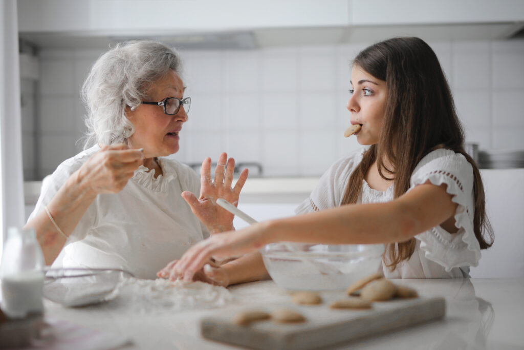 3768146-3768146 Calm senior woman and teenage girl in casual clothes looking at each other and talking while eating cookies and cooking pastry in contemporary kitchen at home